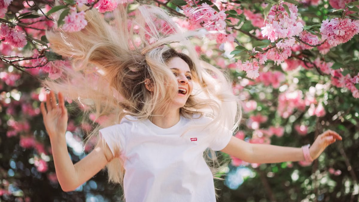 A joyful young woman flipping her hair under blooming pink cherry blossoms on a sunny spring day.